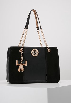 Sac à main noir avec des anses en chaîne dorée, un logo orné de perles et un pendentif nœud beige et noir suspendu, posé sur une surface blanche.