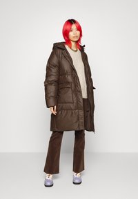 Brun pufferjakke med hætte, der har store frontlommer og lynlås. Båret over en beigefarvet sweater og brune fløjlshorts, med blomsterede sko.