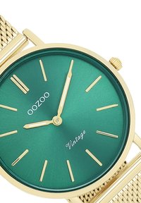 Montre en métal vert avec des accents dorés, dotée d'un cadran minimaliste, de fins marqueurs d'heure et d'un bracelet en maille. Le texte "OOZOO Vintage" apparaît sous le centre.