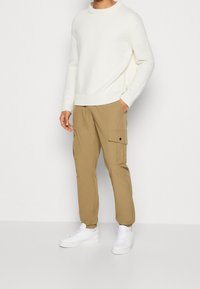 Pull en tricot blanc avec col rond, associé à un pantalon cargo beige avec poches latérales, et porté avec des baskets blanches.