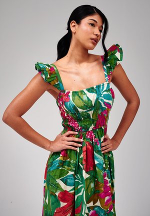 Vrouw met donker haar in een paardenstaart, gekleed in een mouwloze jurk met een bloemenpatroon en ruches op de schouderbanden, staand met handen in de zij.