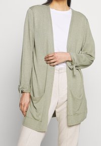Cardigan vert clair à ouverture devant, manches longues, accents noués sur les poignets et deux poches avant. Fabriqué en matériau tricoté doux.