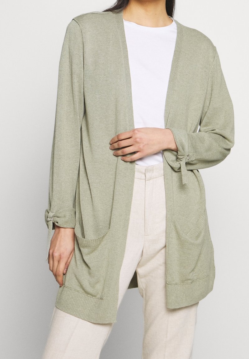 Cardigan vert clair à ouverture devant, manches longues, accents noués sur les poignets et deux poches avant. Fabriqué en matériau tricoté doux.