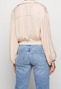 Malli kääntyneenä poispäin, päällään pehmeä beigen värinen paita, jossa on puffipaidan hihat, sekä vaaleansiniset korkeavyötäröiset farkut, esittelee rentoa asua.