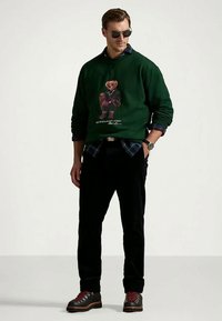 Green sweatshirt with a graphic bear design, layered over a plaid shirt. Black pants and hiking boots complete the outfit. Sunglasses worn.