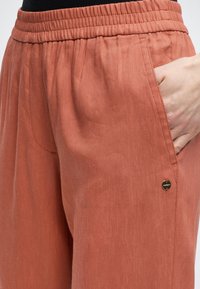 Χέρι που στηρίζεται στην τσέπη χαλαρών, σκουροκόκκινων παντελονιών με ελαστική μέση και μικρό στρογγυλό μεταλλικό λογότυπο.