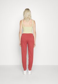 Kvinna med blont hår iförd en ljusgrön linne, röda mjukisbyxor och vita sneakers, stående mot en enfärgad vit bakgrund vänd bort.