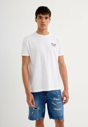Camiseta de algodón blanca con cuello redondo y mangas cortas, que presenta un pequeño logo negro en el lado izquierdo del pecho. Combinada con pantalones cortos de denim desgastados.