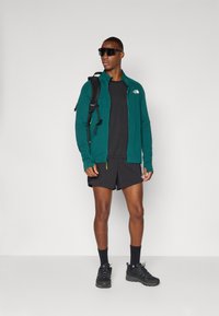 Türkise Zip-Jacke, schwarzes T-Shirt, schwarze Shorts und schwarze Wanderschuhe. Das Modell trägt eine Sonnenbrille und einen Rucksack mit gelben Akzenten.