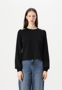 Μαύρη μπλούζα με μακριά μανίκια και χαλαρή εφαρμογή, διακοσμημένη με χάντρες στους ώμους. Συνδυασμένη με μπλε τζιν με ασημένια τρουκ.