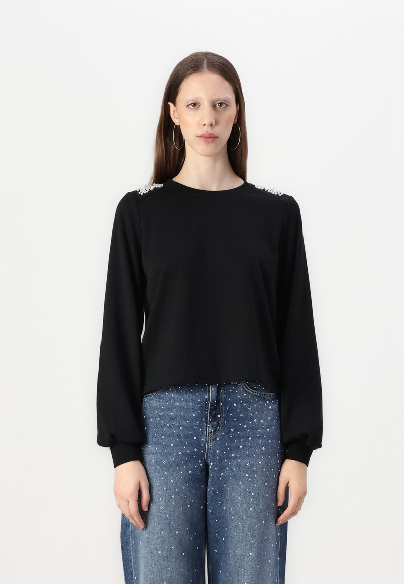 Μαύρη μπλούζα με μακριά μανίκια και χαλαρή εφαρμογή, διακοσμημένη με χάντρες στους ώμους. Συνδυασμένη με μπλε τζιν με ασημένια τρουκ.