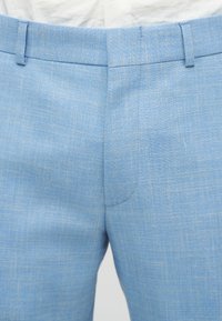 Pantalon bleu clair en tissu texturé avec une finition lisse. Il présente un devant plat et des coutures nettes le long de la ceinture.