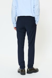 Marinblå byxor med skräddarsydd passform, med en slät textur, två bakfickor med knappar, matchade med en ljusblå skjorta och mörka skor.