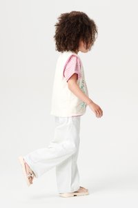 Crème-kleurige vest met groene borduursel over een roze en witte gestreepte shirt, gecombineerd met witte wijde broek en lichte sandalen.