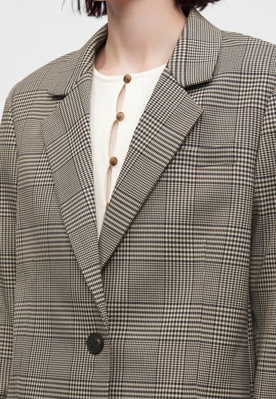 Femme portant un blazer beige et noir à carreaux sur un haut crème côtelé à boutons marron, montrée du cou jusqu'au milieu du torse.