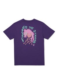 Camiseta de algodón morada con un diseño gráfico en la parte trasera que incluye el texto "Abraza lo Extraño" y detalles ilustrativos en rosa y azul verdoso.