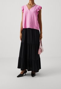 Kvinna i en rosa blus med volanger, svart lager-på-lager-maxikjol, svarta klackar, pärlhalsband och håller en liten rosa handväska.