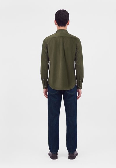 Homme debout de dos, portant une chemise à manches longues vert olive, un jean bleu foncé et des chaussures foncées sur un fond blanc uni.