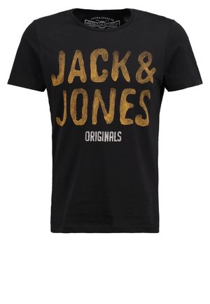 Mężczyzna w czarnej koszulce "Jack & Jones Originals", ciemnych spodniach cargo w kamuflażu, czarnych sneakersach, trzymający na ramieniu pasek czarno-beżowego plecaka.
