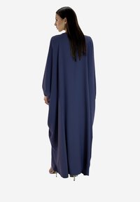 Vrouw die met de rug naar de camera staat, gekleed in een lang, losvallend marineblauw jurk met lange mouwen en hoge hakken, tegen een witte achtergrond.