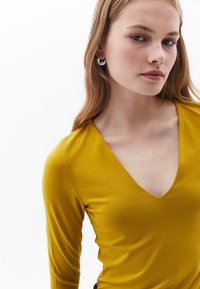 Maglietta gialla a manica lunga con scollatura a V, realizzata in tessuto morbido. Il modello indossa orecchini rotondi in argento. Texture semplice e liscia.