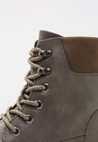Botte en cuir gris avec des accents en daim marron, dotée d'œillets hexagonaux, de lacets tissés et de détails de couture le long de la tige.