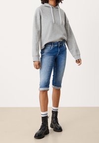 Šedá mikina s kapucí a bílými stahovacími šňůrkami, modré džíny bermudy, černé šněrovací kotníkové boty a bílé ponožky s černými pruhy.