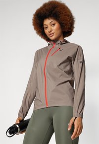 Letvægts, taupe løbejacket med en rød lynlås og hætte, som har elastiske manchetter og en lynlås brystlomme, parret med grønne leggings.