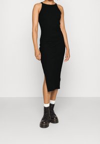 Femme portant une robe midi noire sans manches et côtelée avec une fente sur le côté, assortie de chaussettes blanches et de bottes de combat noires à lacets, se tenant sur un sol gris.