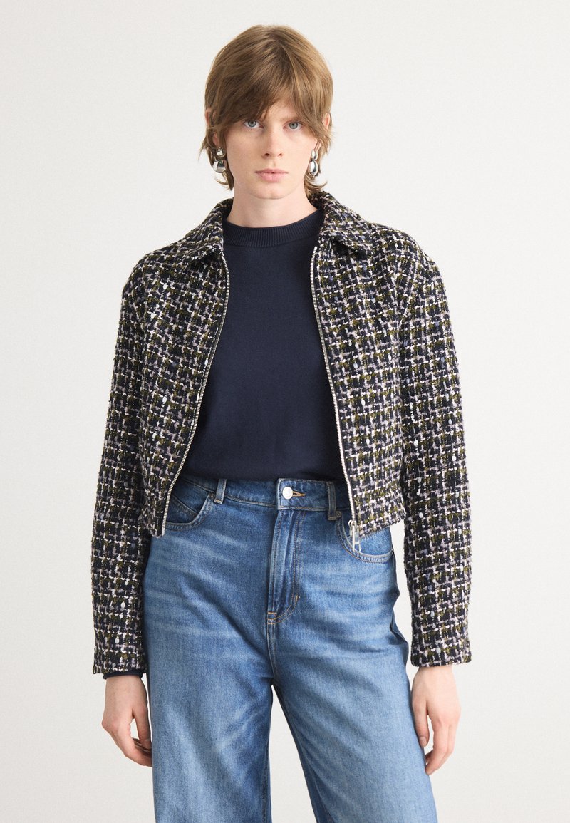 Giacca cropped bicolor con un motivo checkered testurizzato nei colori blu navy, bianco e verde. Presenta un colletto classico e una zip argentata. Indossata sopra un top scuro.