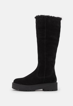 Botte haute jusqu'au genou en daim noir avec semelle épaisse à crampons et bordure en fausse fourrure en haut, présentée sur un fond blanc uni.