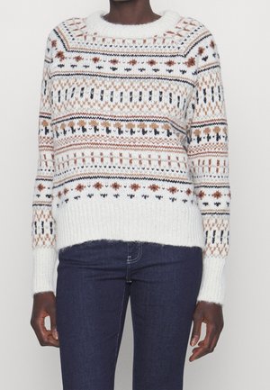 Osoba w białym swetrze z dzianiny z wielokolorowym geometrycznym wzorem i dopasowanych ciemnoniebieskich dżinsach, stojąca na jednolitym tle.