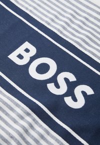 Marineblått stoff med hvite striper og med fet tekst "BOSS" i hvitt. Materialet ser teksturert ut, noe som fremhever designdetaljene.