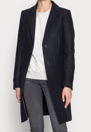 Kvinde iført en sort mellemlang frakke over en lysegrå sweater og mørkegrå jeans mod en ensfarvet baggrund.