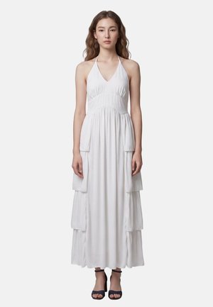 Femme portant une robe maxi blanche à col halter avec des couches superposées et des talons noirs ouverts à l\'avant, debout devant un fond uni.