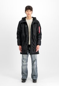 Melnā auduma parka ar kapuci, ar divām priekšējām kabatām, ar rāvējslēdzēju un sarkanu birku. Uzvilkts virs gaiši bēša krekla un izbalējušām zila džinsu biksēm.