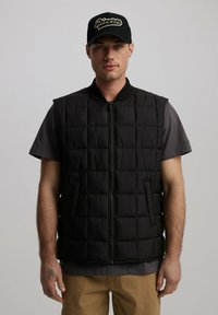 Czarna pikowana kamizelka z wysokim kołnierzem i zamkiem błyskawicznym z przodu. Posiada dwie boczne kieszenie. Nosi się ją na szarej koszulce, w zestawieniu z khaki spodniami.