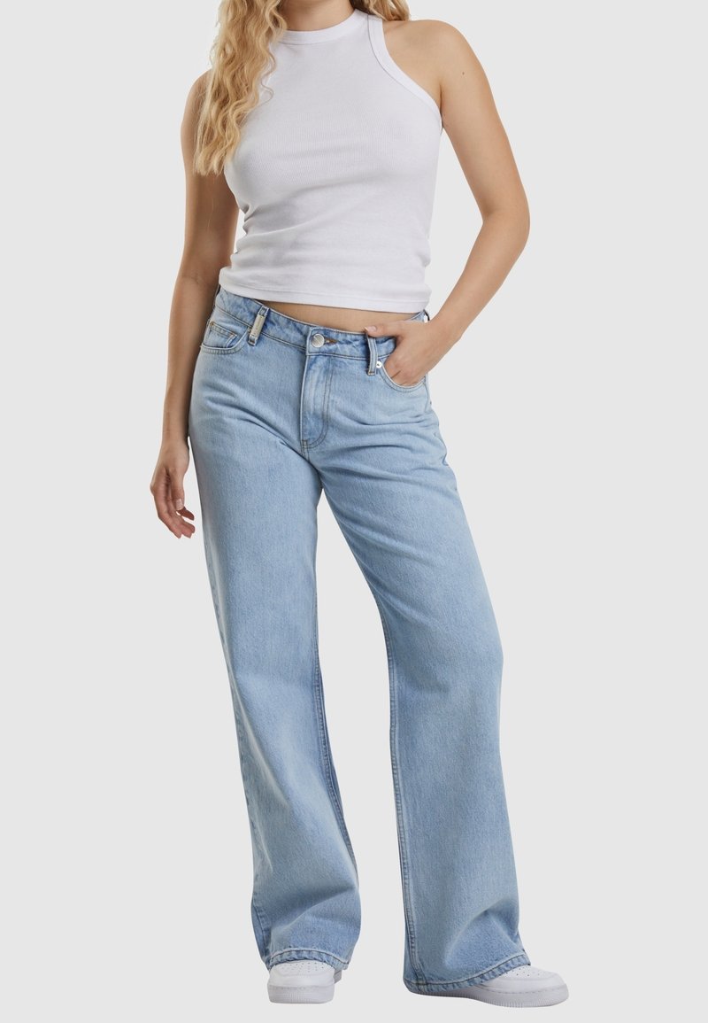 Femme portant un haut court blanc sans manches, un jean large bleu clair et des baskets blanches, debout avec une main dans la poche.