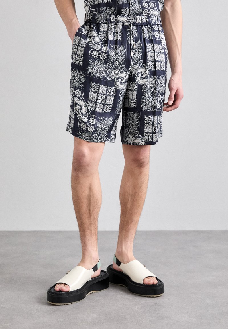 Dark blue patterned shorts with elastic waistband and pockets; intricate white floral and paisley designs. Model wears chunky white sandals.