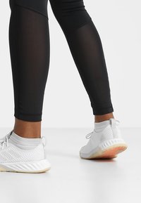 En person som bär vita löparskor och svarta leggings står på en vit yta mot en enfärgad vit bakgrund.