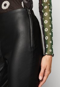 Gros plan d’une main près d’un pantalon en cuir noir avec fermeture éclair latérale, accompagné d’un haut transparent à manches longues avec des motifs floraux blancs sur tissu vert et noir.