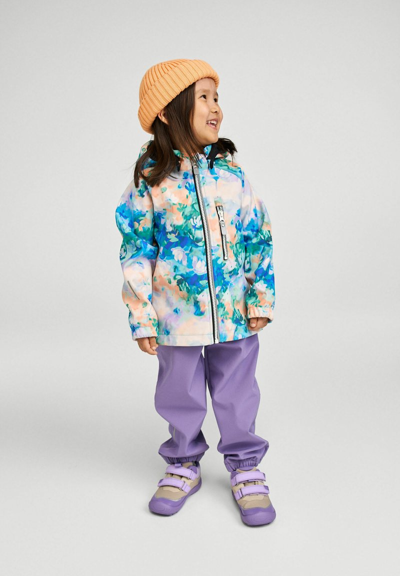 Małe dziecko w kolorowej kurtce w kwiaty, lawendowych spodniach, fioletowych trampkach i pomarańczowej dzianinowej czapce, uśmiechające się i patrzące na bok.