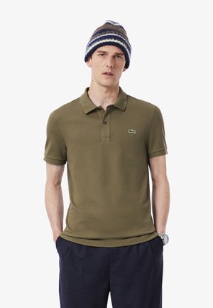 Mand iført grøn polo med logo, marineblå bukser, stribet strikhue og sølvfarvet armbåndsur, stående med hænderne i lommerne.