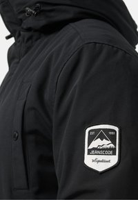 Zwarte geïsoleerde jas met een capuchon, met een patch op de mouw die bergen weergeeft, merknamen "JEANSCODE," en opgericht in 1993.