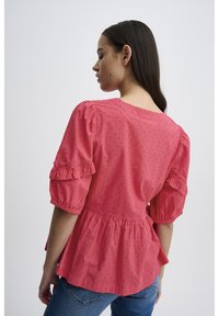 Blouse rose avec des manches courtes à volants et un ourlet peplum. Présente un motif texturé et un décolleté arrière arrondi. Portée avec un jean bleu.