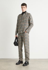 Mintás szövet szett, amely egy galléros ingből és egy hozzá illő nadrágból áll fekete, narancs és krém színekben. A modell egy fekete táskát tart és fekete cipőt visel.