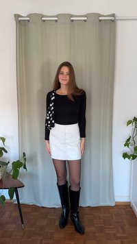 Fekete hosszú ujjú felső, fehér miniszoknya elülső zsebekkel, fekete térdig érő csizma és pöttyös válltáska. Beltéri környezet növényekkel.