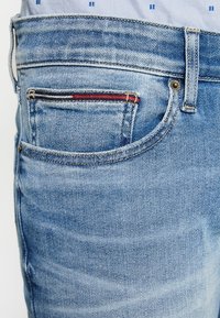 Vybledlé modré džíny s designem pěti kapes, které obsahují malou kapsu s červenými a námořnickými akcenty. Texturovaný denimový povrch.