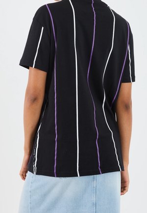 Personne portant une chemise noire à manches courtes avec des rayures verticales blanches et violettes, associée à une jupe en jean bleu clair, vue de dos.