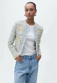 Giacca cropped multicolore con texture, caratterizzata da un bordo frangiato, due tasche frontali, indossata sopra una camicia bianca e jeans azzurri chiari.